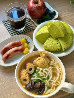 Overhead view of a meal with noodle soup, sausages, green cake slices, apple, blueberries, and a drink