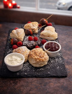 Slate board with scones, fresh berries, jam bowl, and powdered sugar on wooden table