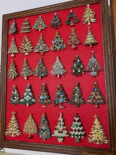 Framed display of approximately 30 decorative Christmas tree brooches on red velvet background