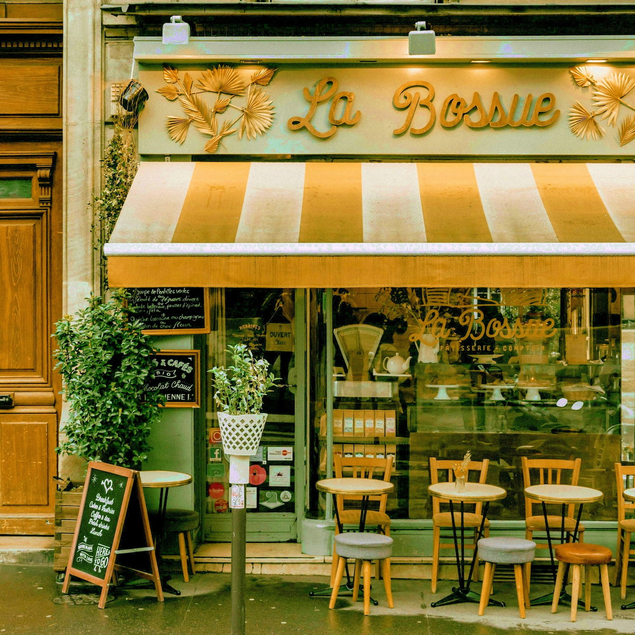 French cafe storefront with striped awning, outdoor seating, chalkboard sign, and potted plants
