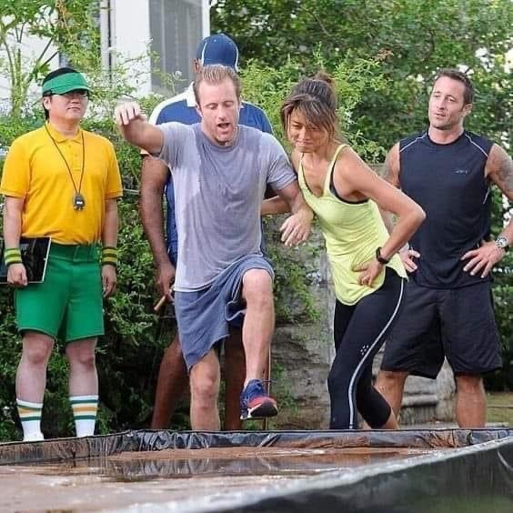 Four adults crossing a low obstacle outdoors near bushes and a building