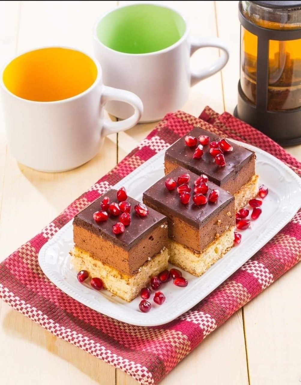 Three cake squares with chocolate topping and pomegranate seeds on white plate with colorful mugs
