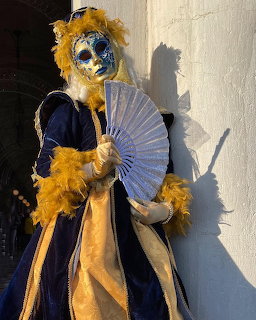 Person in ornate blue and gold Venetian carnival mask and costume with feathered headdress holding a fan