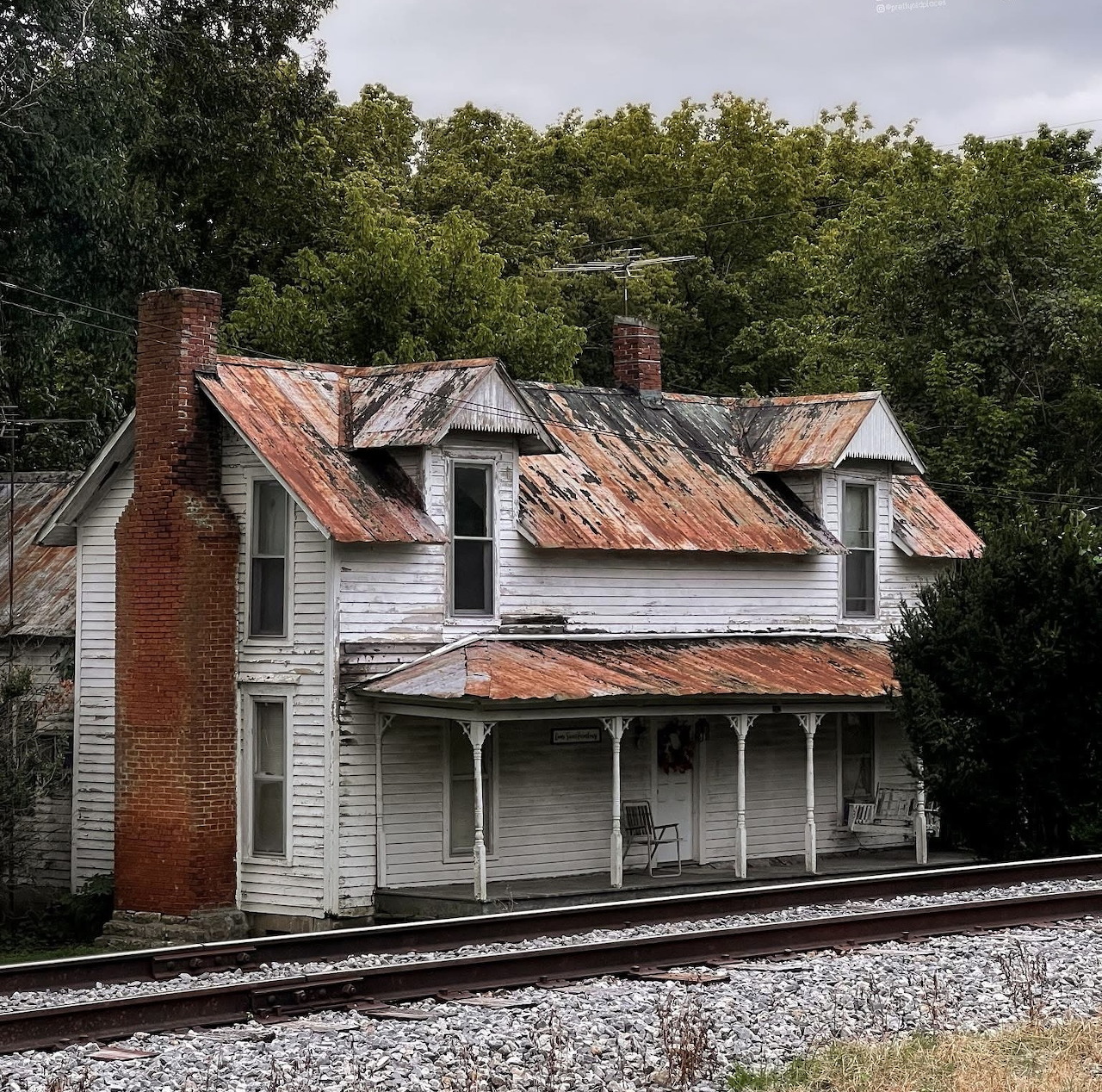 Old white wooden house with a rusty roof beside railroad tracks and trees