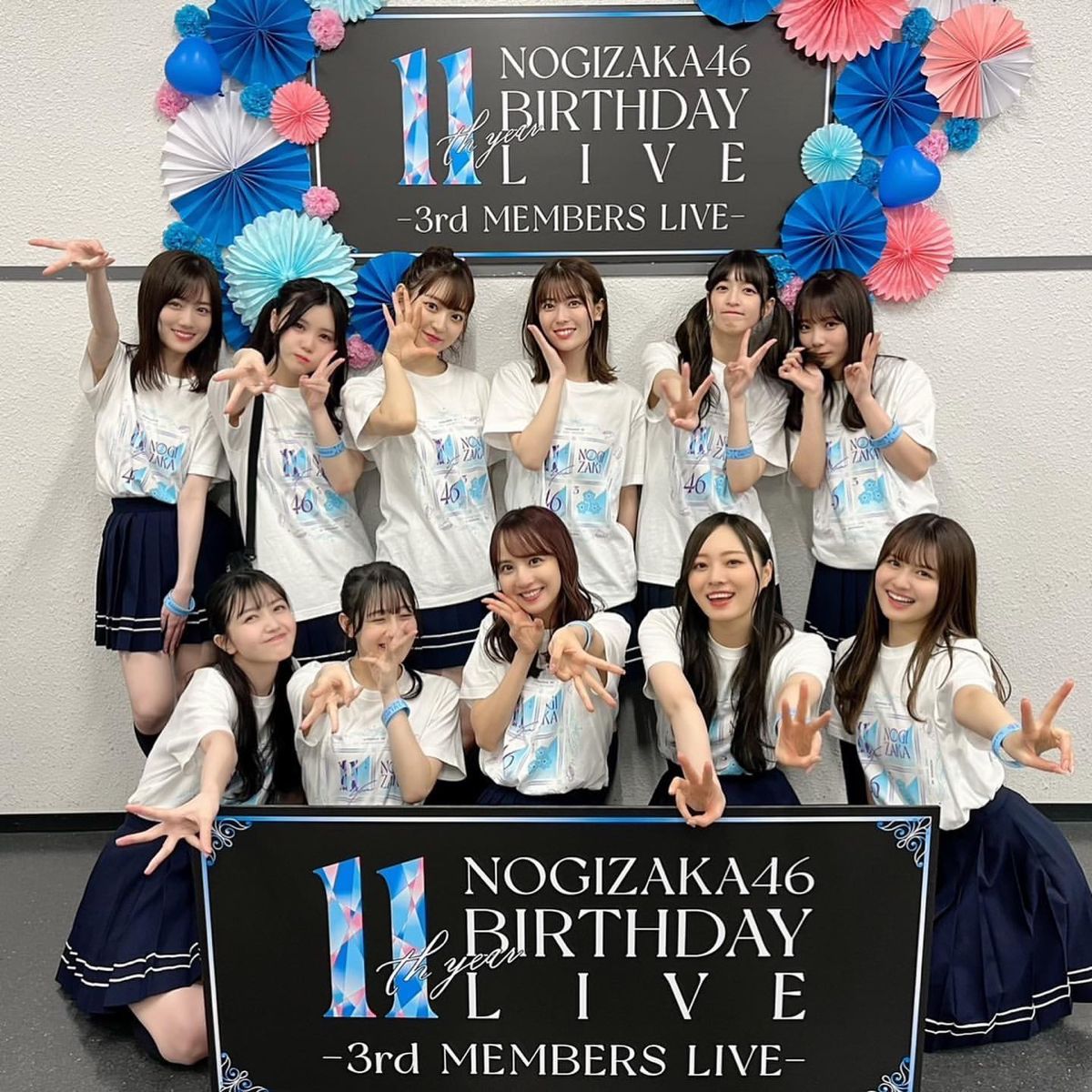 Eleven women posing in matching outfits in front of a Nogizaka46 Birthday Live event sign