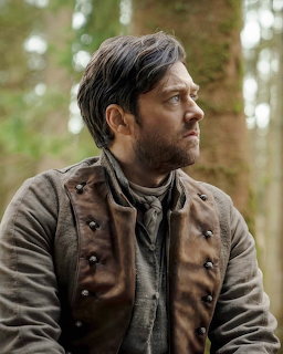 Man in period brown coat with buttons and grey cravat standing in a woodland setting