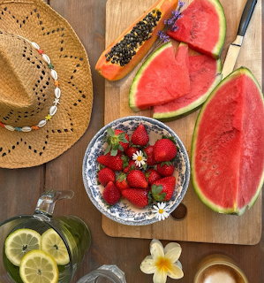 Flatlay of watermelon, papaya, strawberries in bowl, lemon water, and straw hat on wooden surface