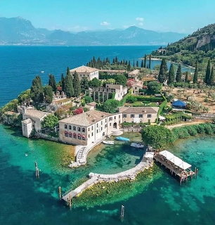 Villa complex on a narrow peninsula in a turquoise lake with cypress trees and distant mountains