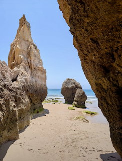 Tall rock formations on a sandy beach framed by rocky overhang with ocean and blue sky behind