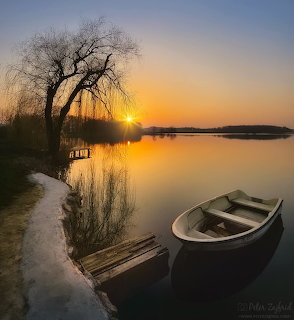 Small boat on a lakeshore at sunset with a bare tree and wooden dock reflected in calm water