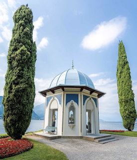 White pavilion with dome between two cypress trees on Lake Como shore with mountains and blue sky