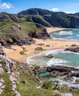 Sandy beach cove surrounded by rocky cliffs and green hills with ocean waves and blue sky
