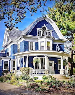 Blue Victorian-style house with white trim, wraparound porch, and gambrel roof surrounded by trees