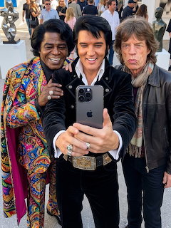 Three celebrity figures posing for a selfie outdoors, with Elvis Presley centered between James Brown and Mick Jagger.