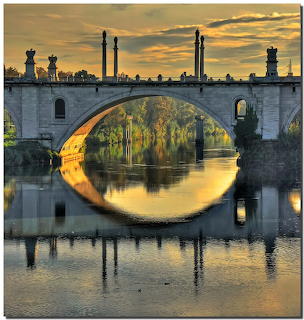 Stone bridge with decorative pillars reflecting in still water at sunset with golden sky