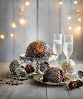 Christmas puddings on a cake stand with pine cones, ornaments, and champagne glasses against bokeh lights