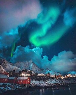 Aurora borealis glowing green and blue over snow-covered mountains and coastal buildings at night