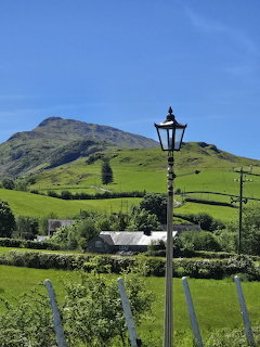 Railway platform with lamp post overlooking green mountain and fields under blue sky