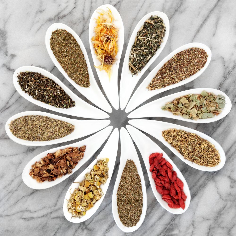 White spoons arranged in circle containing different dried tea leaves and herbs on marble background