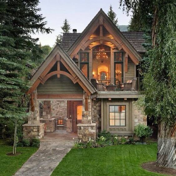 Stone and wood cottage with lit windows at dusk, surrounded by evergreen trees and green lawn