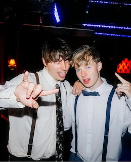 Two young men in white shirts and suspenders posing together in a dark club or party setting.