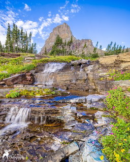 Waterfall cascading over layered rocks with a tall rocky mountain peak and evergreen trees in the background