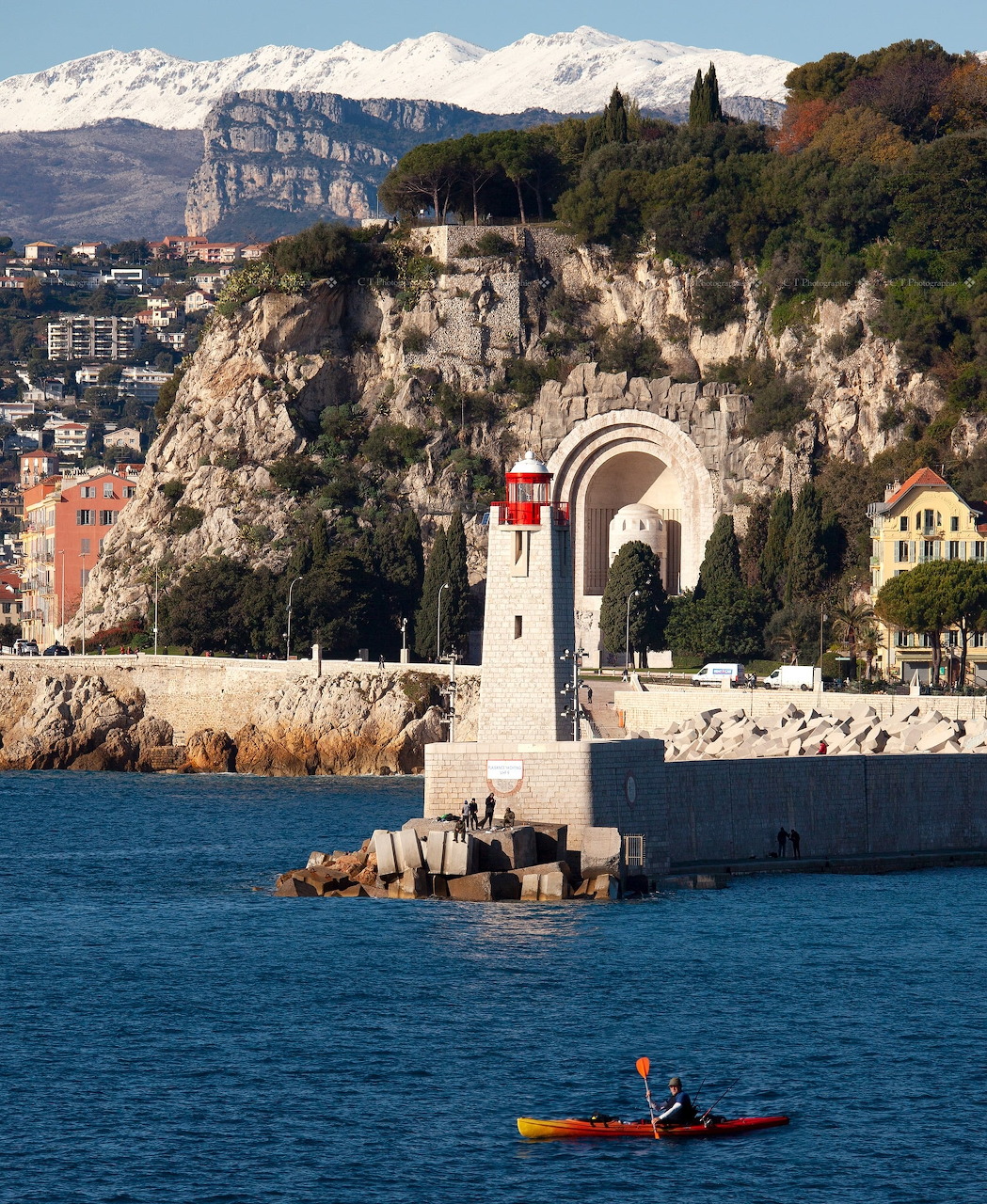 Kayaker near a white lighthouse and arch monument at Antibes harbor with snowy mountains behind