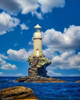 Stone lighthouse standing on a narrow rocky island surrounded by blue sea under cloudy sky