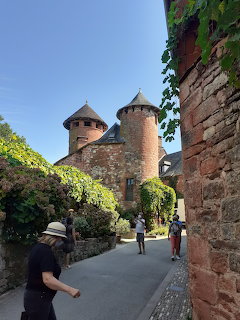 Stone castle towers above a narrow walkway with old walls, greenery, and a few people