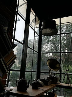 Dark cafe interior with tall windows, rain outside, a wooden table, books, and hanging lamps