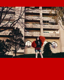 Young woman in red oversized top posing outdoors in front of a multi-story building with trees
