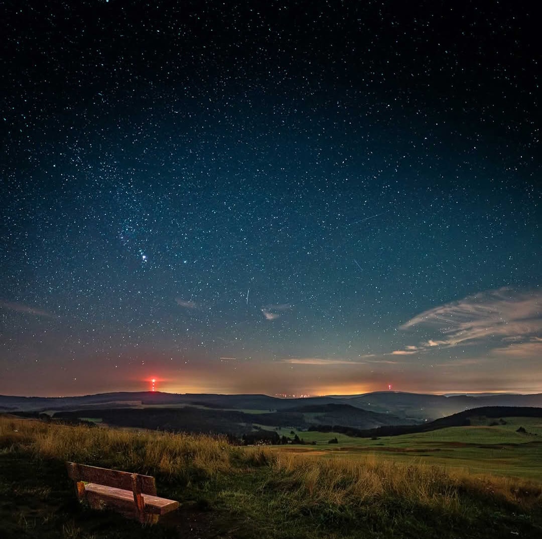 Starry night sky over rolling hills with a wooden bench in the foreground