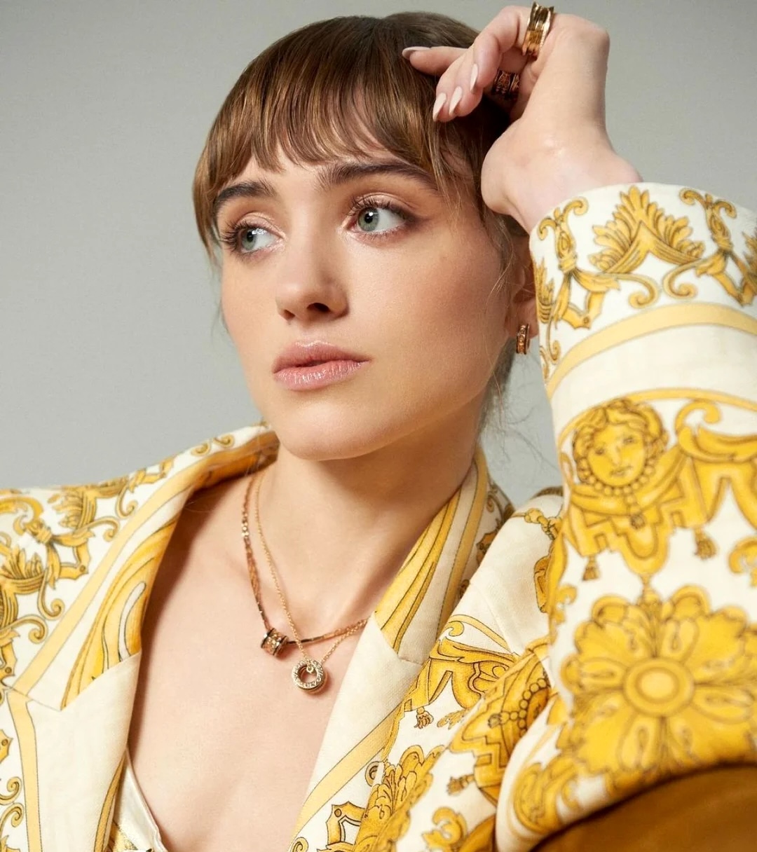 Young woman with bangs in a gold and white patterned blazer posing with hand near her head