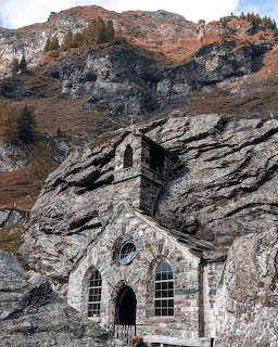 White chapel with three arched windows carved into gray rocky cliff beneath mountain