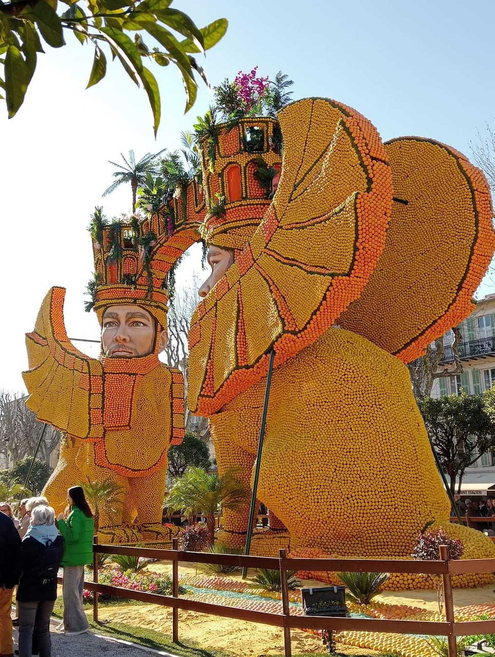 Giant orange citrus fruit sculptures of human figures at outdoor festival with Mediterranean architecture background