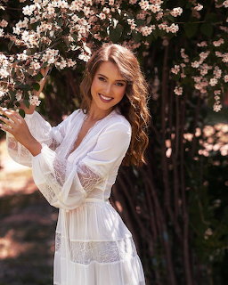 Smiling woman in white lace robe reaching for pink cherry blossom branches outdoors