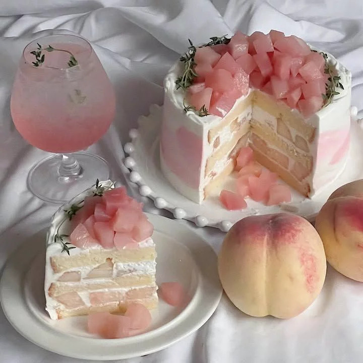 Peach layer cake with a slice, pink drink, and fresh peaches on white fabric