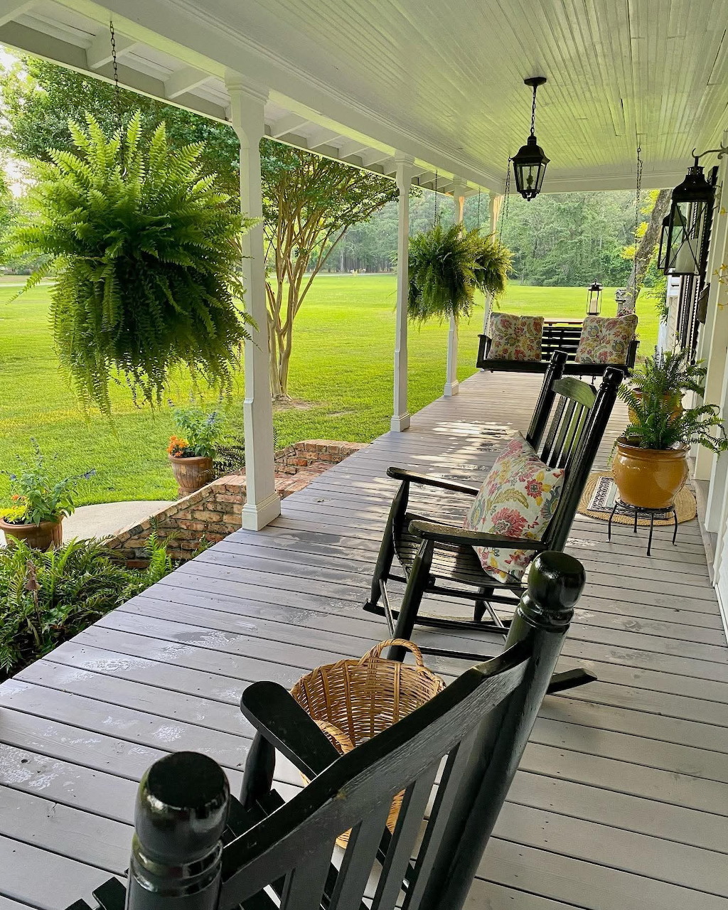 Covered porch with rocking chairs, hanging ferns, potted plants, and a grassy yard