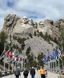 Mount Rushmore sculpture with four presidents carved in mountain face, flags and visitors on walkway below