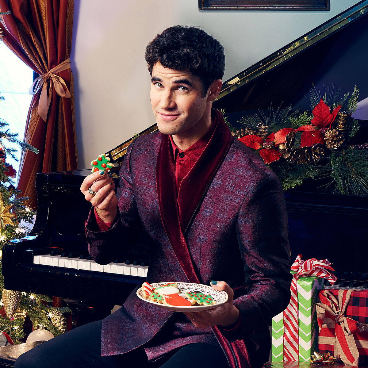 Man in burgundy suit holding cookie and plate with Christmas wreath and piano behind him