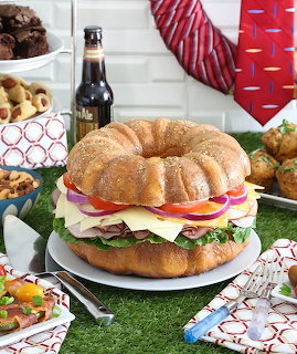 Large ring-shaped sandwich with layers of meat, cheese, and vegetables on a decorated party table with a red tie