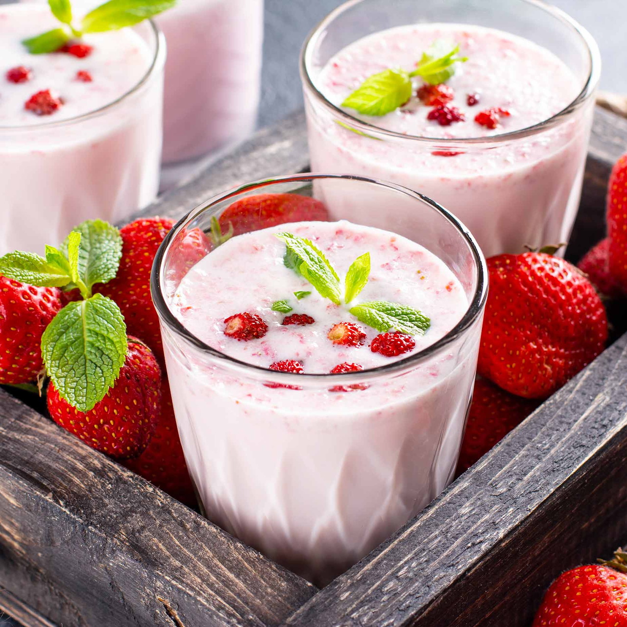 Three glasses of pink strawberry yogurt drinks with mint garnish and fresh strawberries on wooden tray