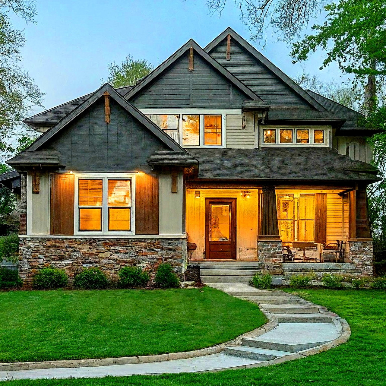 Two-story craftsman style house with stone accents, illuminated windows, curved front walkway and green lawn at dusk