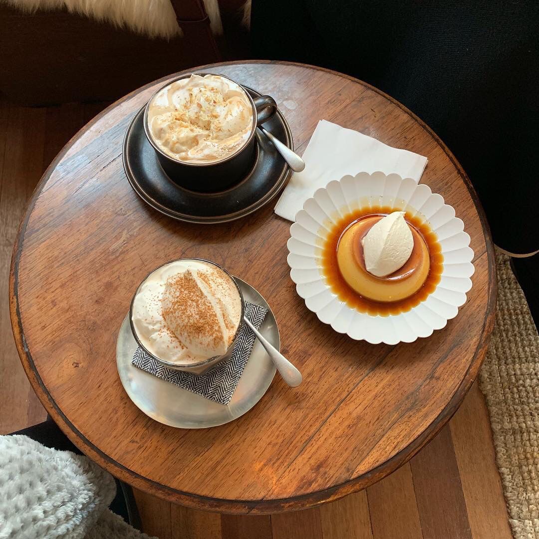 Top-down view of two coffee drinks and a caramel pudding on a round wooden cafe table