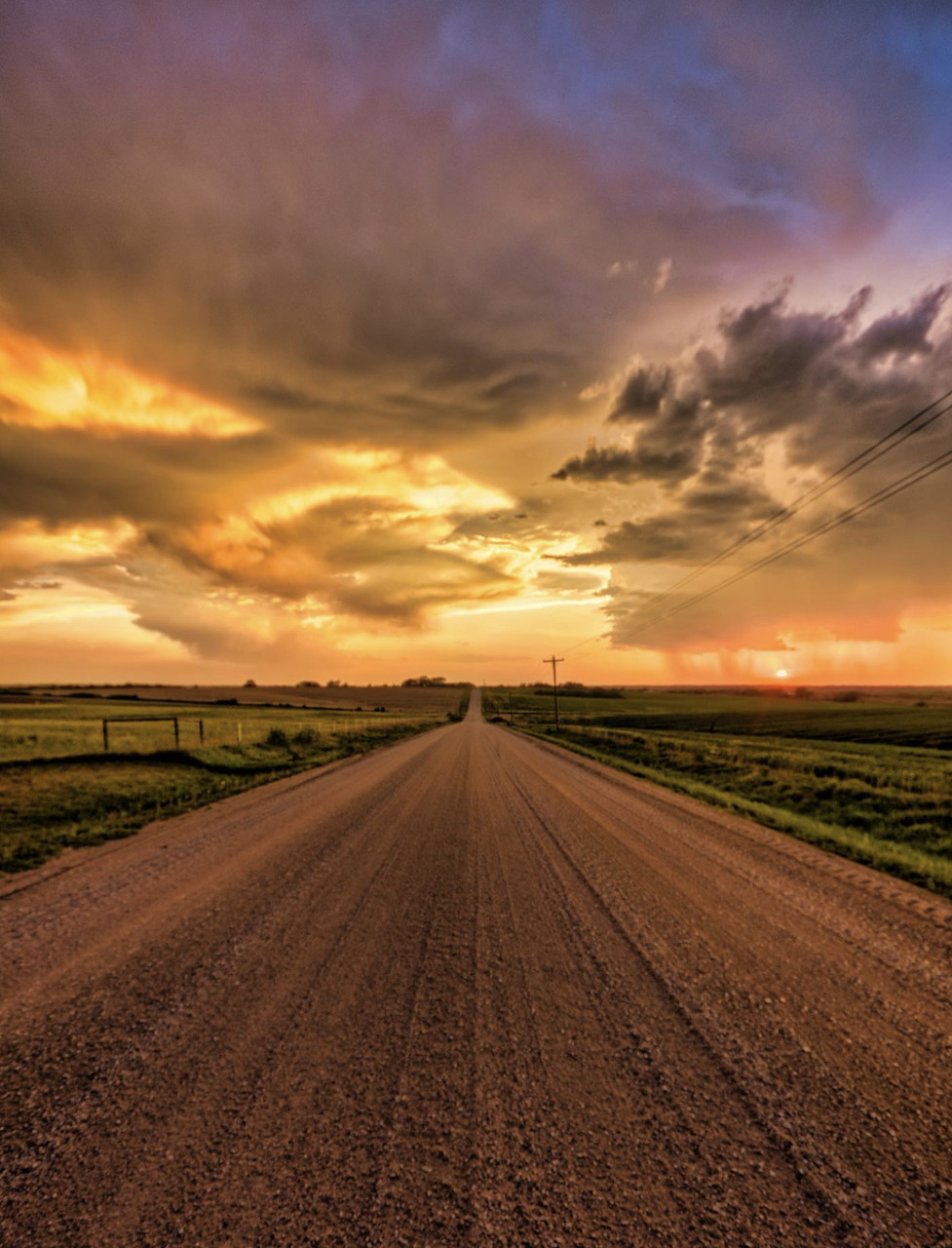 Gravel country road leading to horizon under colorful sunset sky with clouds over farmland