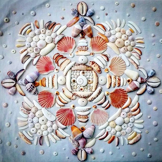 Symmetrical mandala made from white and coral seashells, stones, and shells arranged in circular pattern on pale blue surface