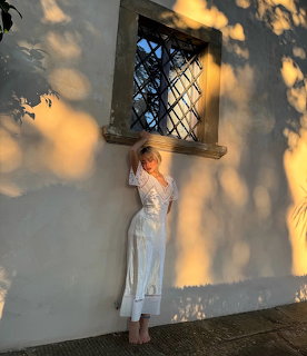 Woman in a white dress standing by a sunlit wall under a small barred window
