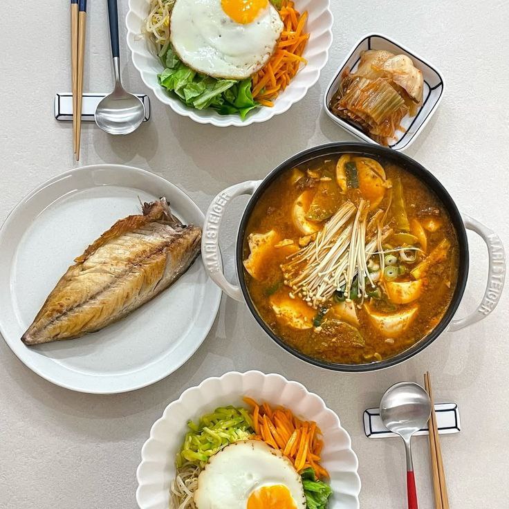 Top-down Korean meal with stew, egg rice bowls, grilled fish, and kimchi on a white table