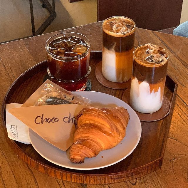 Three iced coffee beverages with a croissant on a wooden serving tray