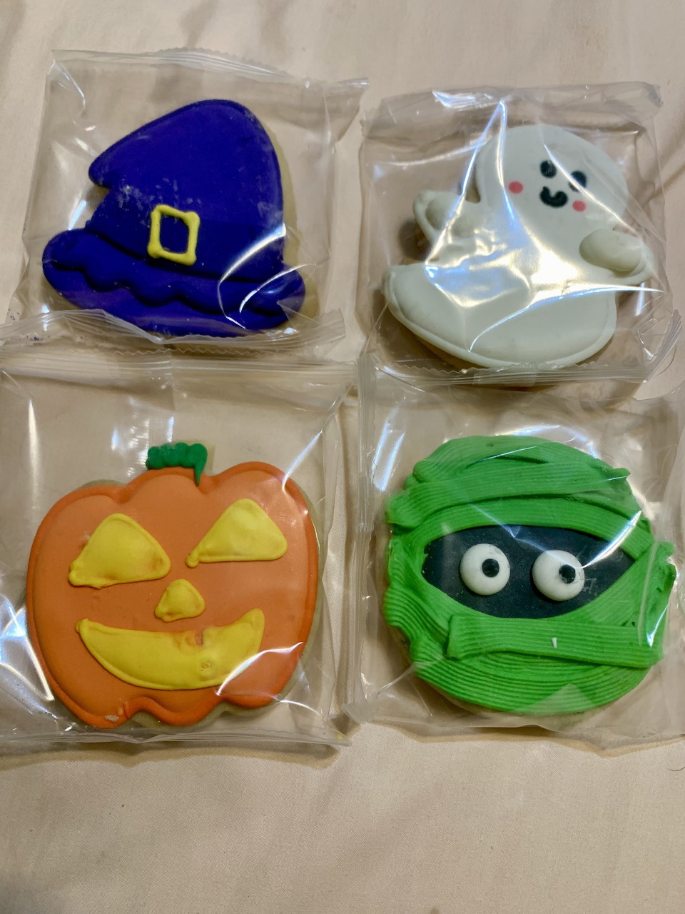 Four Halloween cookies: witch hat, ghost, pumpkin, and mummy in clear wrappers on white cloth
